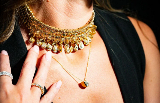 Gold diamond choker necklace with dangling gemstones worn on neck, paired with layered gold pendant and diamond rings.